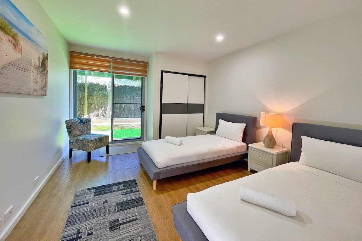 Un dormitorio moderno con cama, mesitas de noche, una silla y vistas a un jardín por un ventanal corredero.