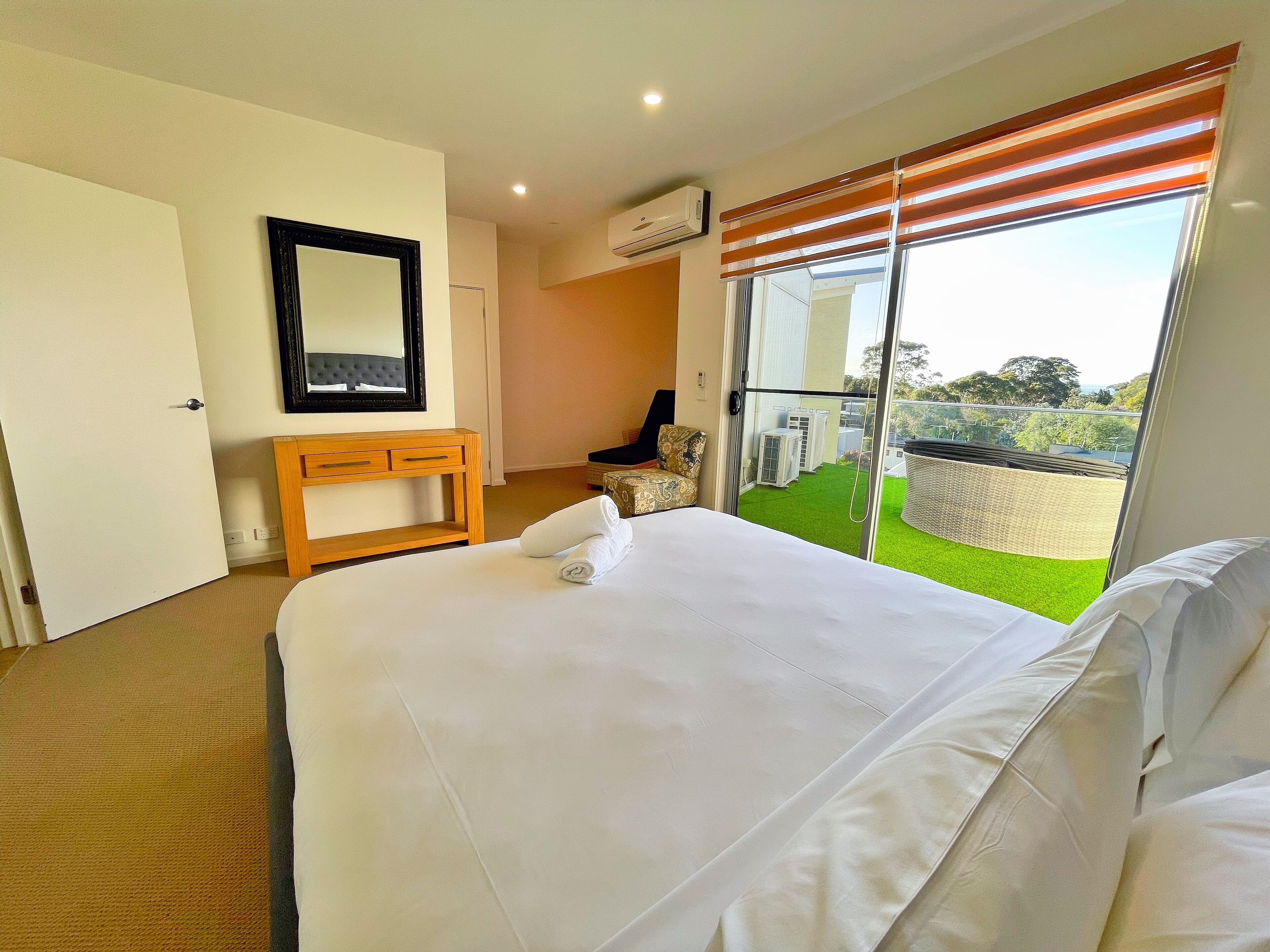 A hotel room with a large bed, a bedside table, a mirror, and a view of a green lawn and air conditioning units outside.