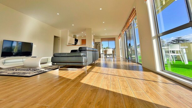 A modern living room with a flat-screen TV, grey sofa, and a large window offering a view of the outdoors. - Phillip Island Towers (Cowes)