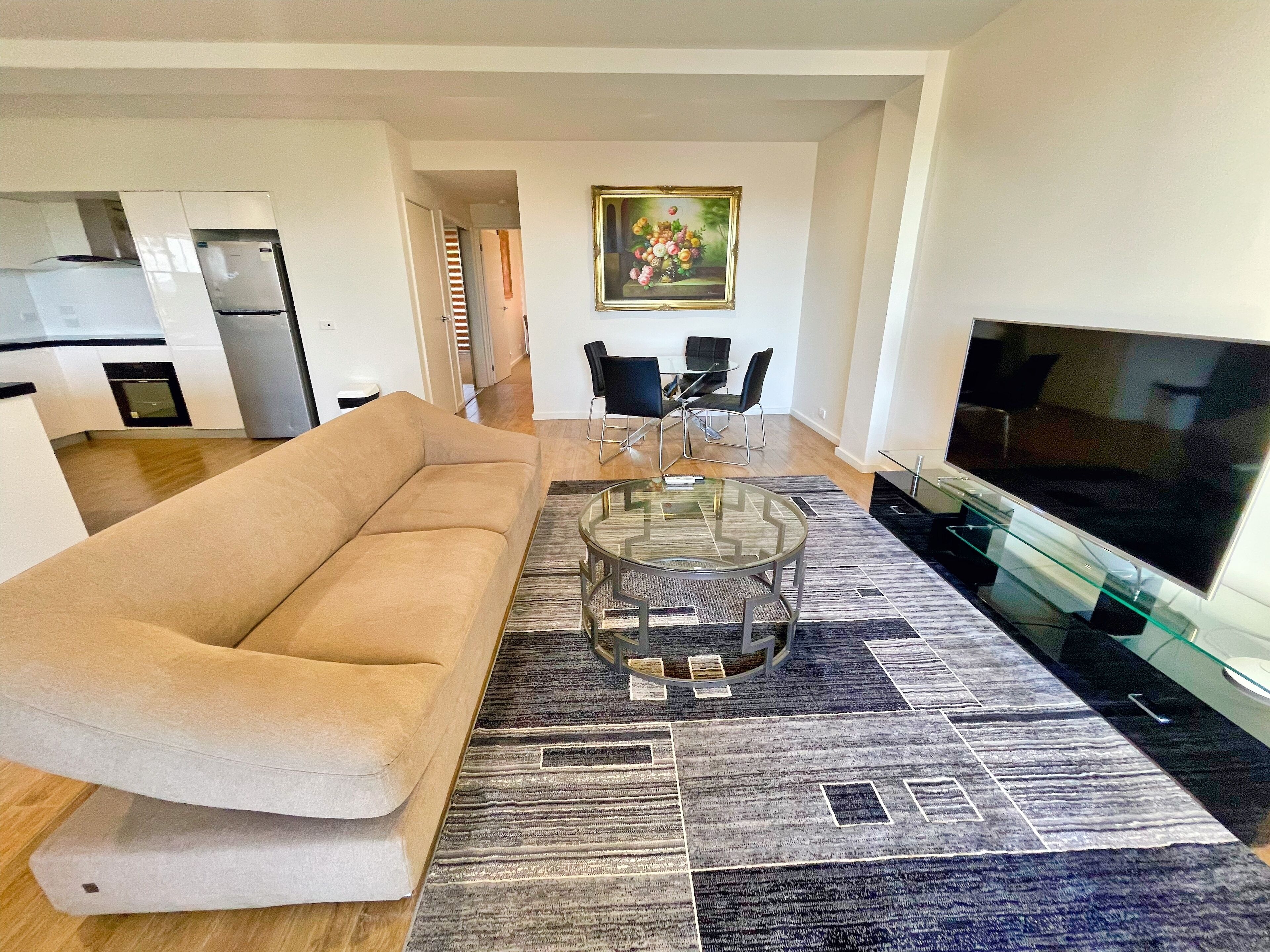 A modern living room with a beige sofa, a glass coffee table, a flat-screen TV, and a patterned rug.