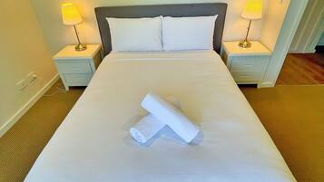 A neatly made bed with white linens, two pillows, and a rolled towel on the bed. There are two bedside lamps on either side of the bed.