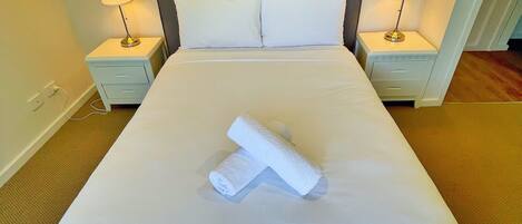 A neatly made bed with white linens, two pillows, and a rolled towel on the bed. There are two bedside lamps on either side of the bed.