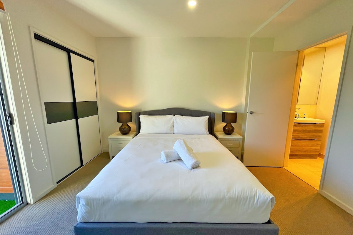 Una cama bien hecha con sábanas blancas y una toalla enrollada, dos lámparas de noche, un cabecero oscuro y una puerta corredera que da a un baño.