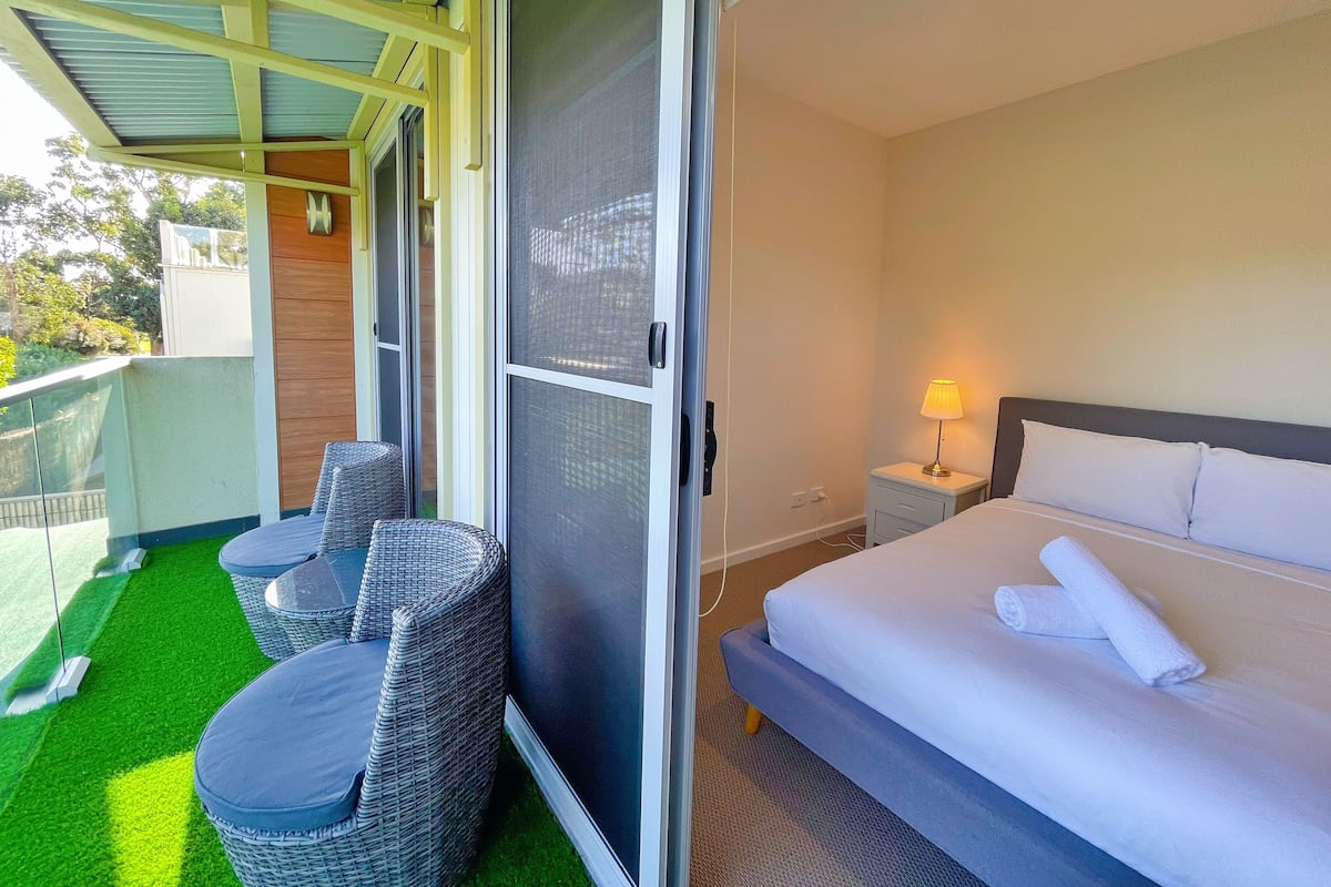 Habitación de hotel con cama, una mesita con lámpara, una silla y un balcón con césped artificial y mobiliario de mimbre.