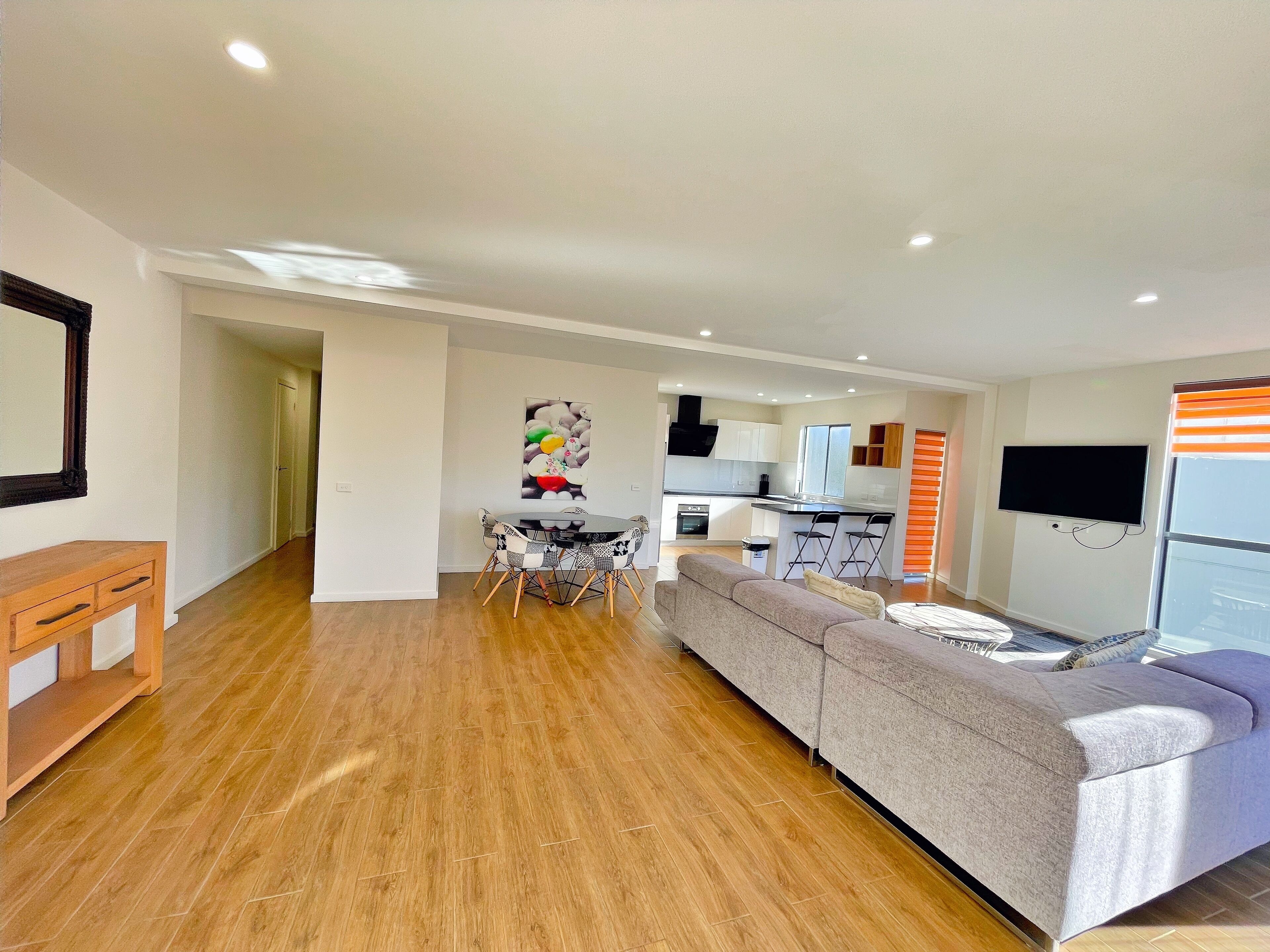 A modern living room with a grey sofa, a dining area with a round table, and a kitchen with white cabinets and a black countertop.
