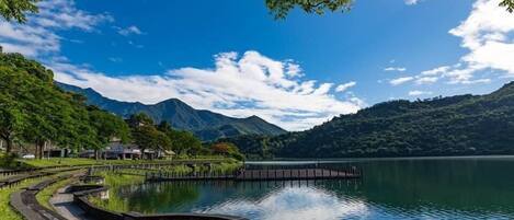 Un parque tranquilo junto al lago con una pasarela de madera, cielo azul despejado y montañas al fondo.