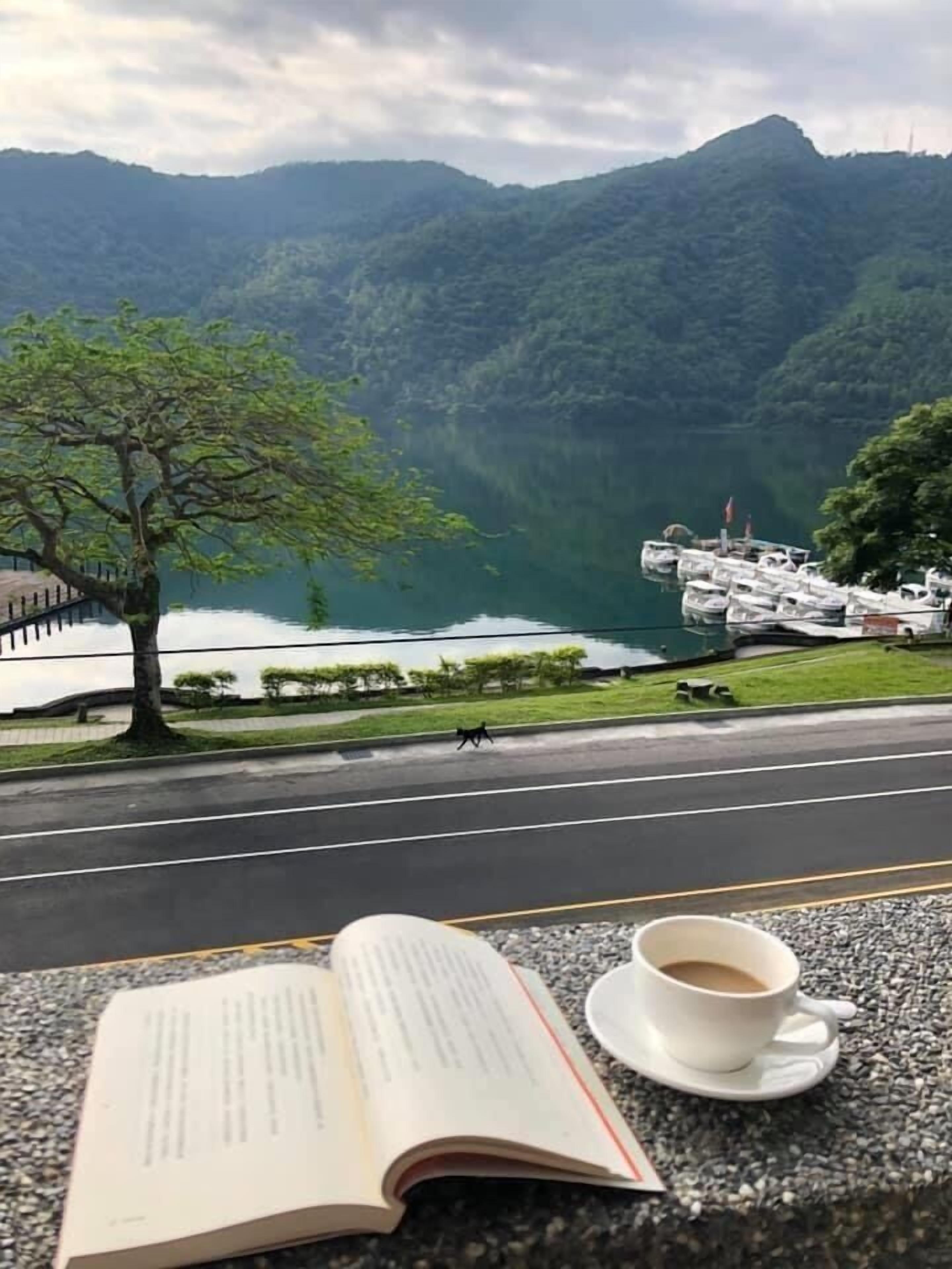 A book and a cup on a table with a view of a lake and mountains.