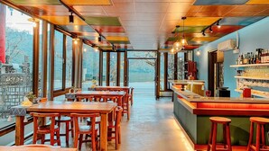 Un restaurante con mesas y sillas de madera, una zona de bar y grandes ventanales con vistas al exterior.