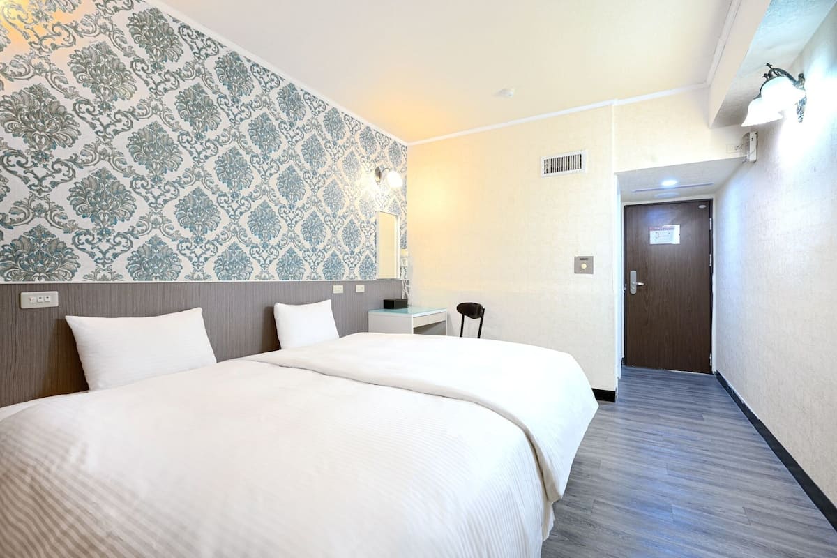 A hotel room with two beds, a small desk, a chair, and a bathroom visible through an open door.