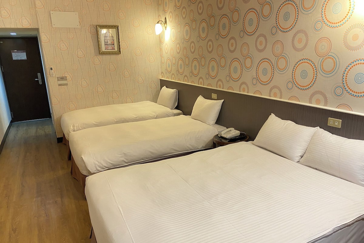 A hotel room with two single beds and a larger bed, a bedside telephone, and a wall-mounted light fixture.