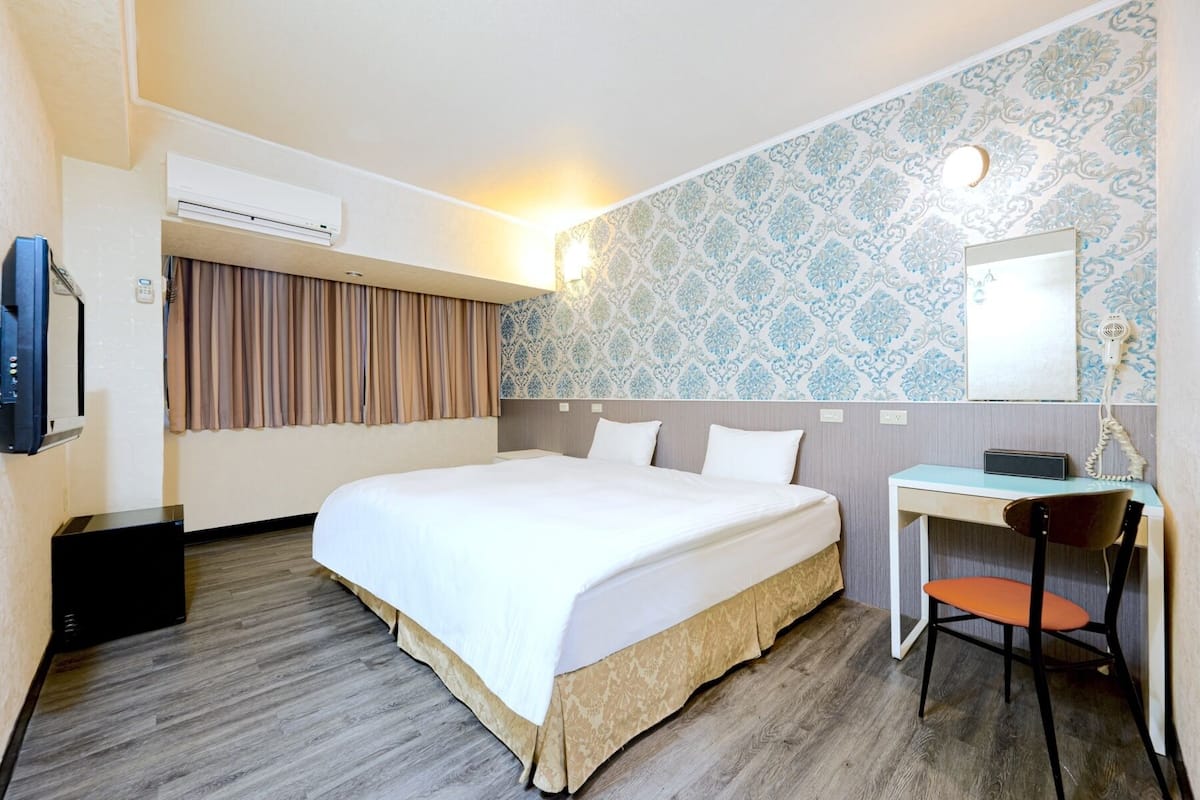 A hotel room with a large bed, a desk, a chair, an air conditioning unit, and a patterned wallpaper.