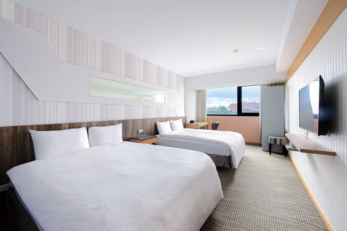 A hotel room with two beds, a TV, a desk, and a window with a city view.