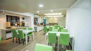A modern dining area with green chairs and white tables, a kitchen area with a counter and appliances, and a view of the city through large windows. - Sunny Hotel (Taipei)