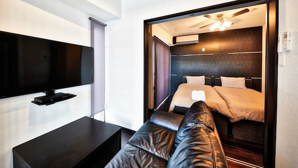 A modern hotel room with a flat-screen TV mounted on the wall, a dark wooden side table, and a large bed with a headboard. - Terrace Resort Shintoshin (Naha)