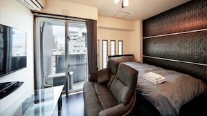 A modern hotel room with a sofa, a bed with neatly folded towels, a flat-screen TV, and a view of the city through a large window. - Terrace Resort Shintoshin (Naha)
