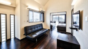 A modern hotel room with a black leather sofa, a wooden coffee table, a bed with a city view, and a large mirror. - Terrace Resort Shintoshin (Naha)