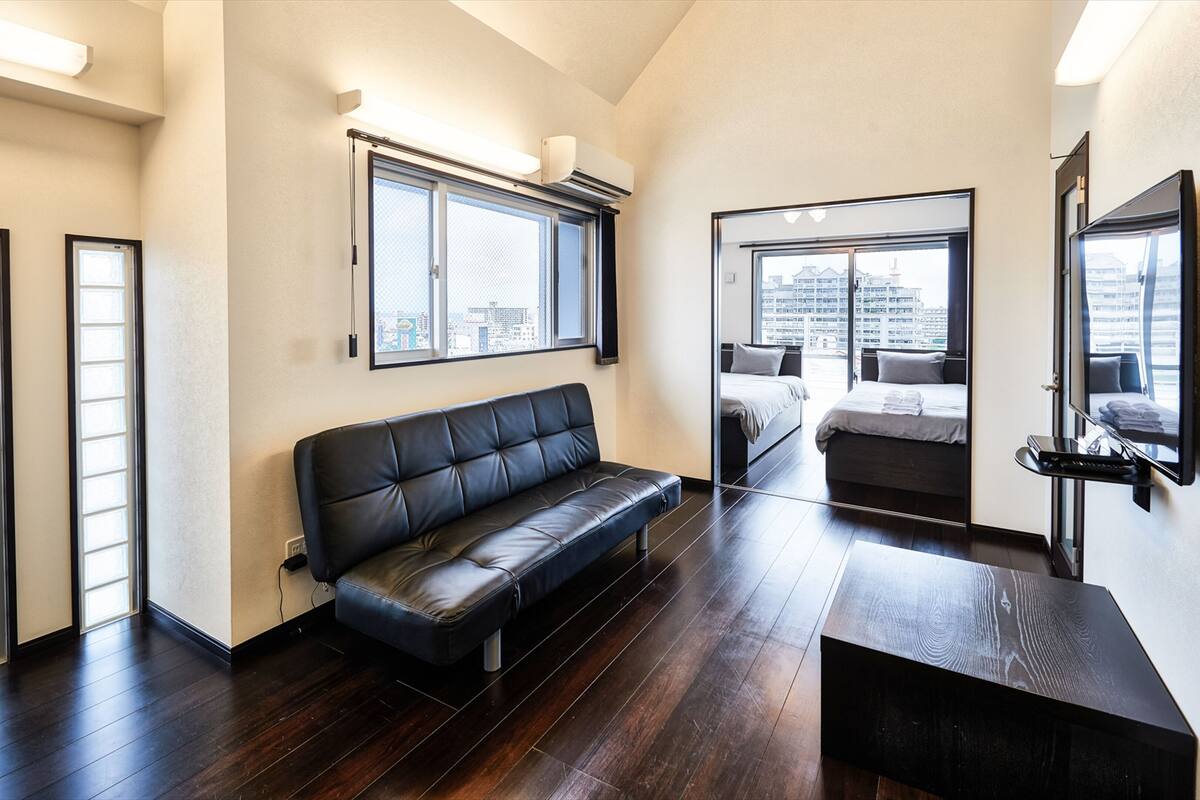 Une chambre d’hôtel moderne avec un canapé en cuir noir, une table basse en bois, un lit donnant sur la ville et un grand miroir.