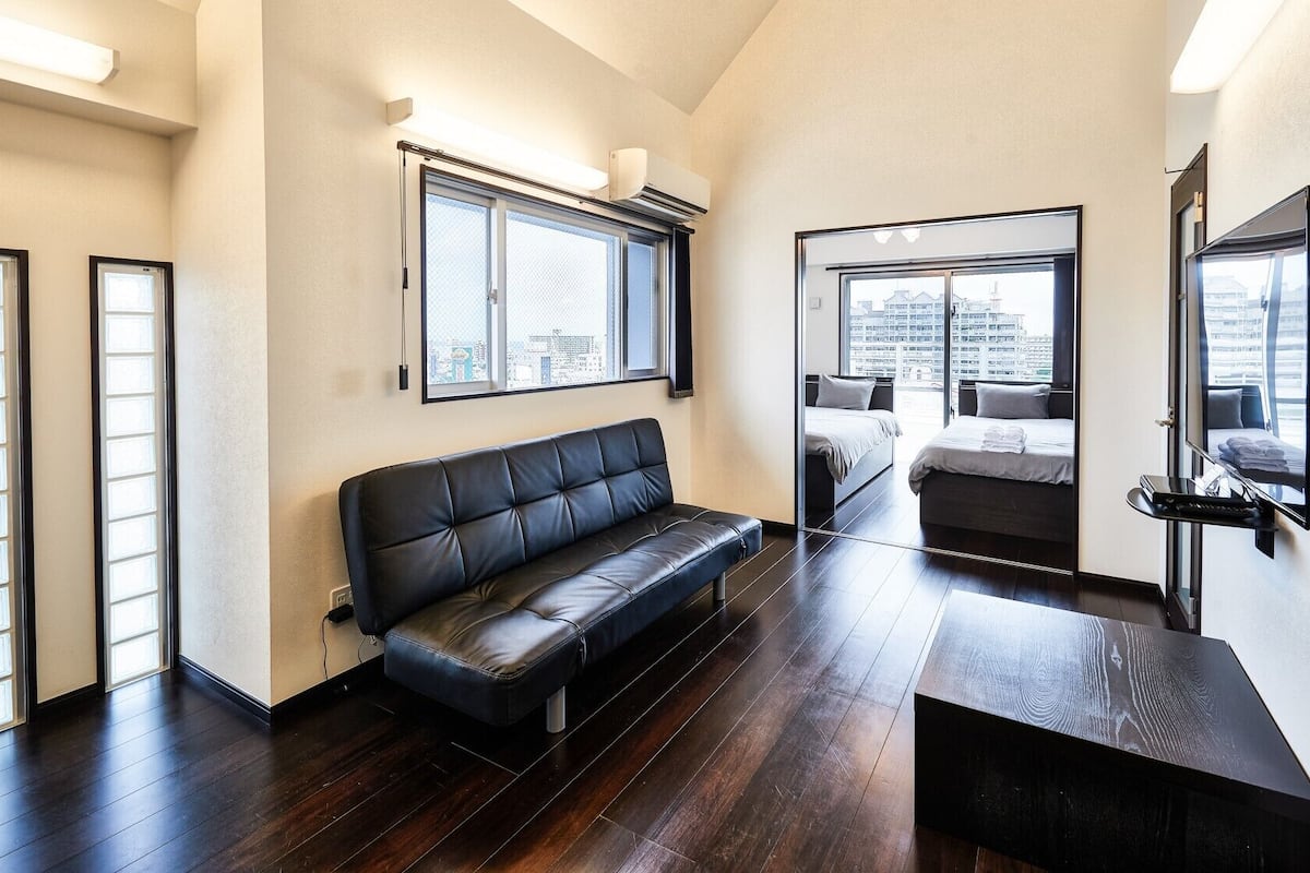 Quarto de hotel moderno com sofá de couro, cama, TV e grandes janelas com vista para a cidade.