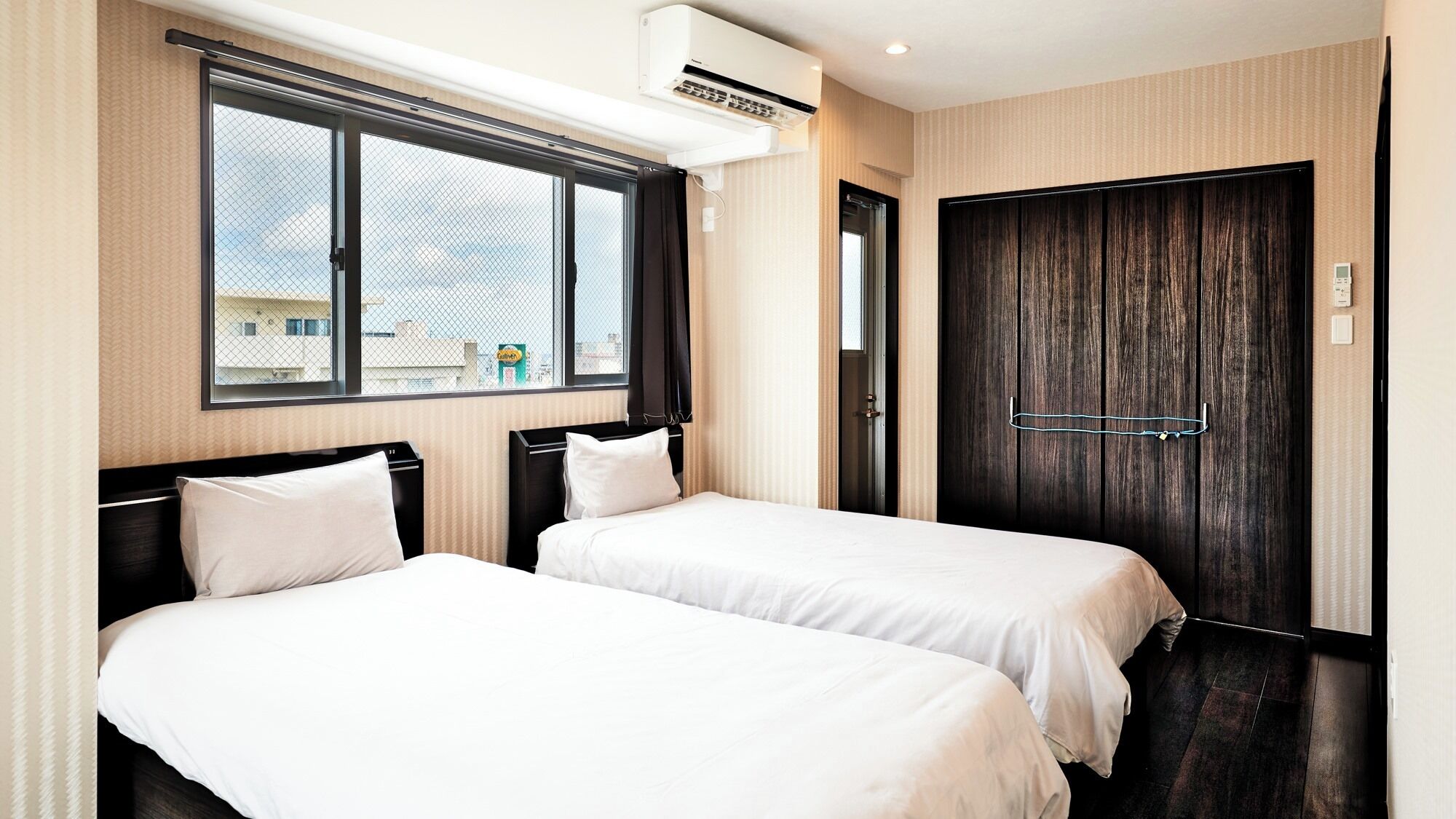 Ein Hotelzimmer mit zwei Betten, einem großen Fenster, einer Klimaanlage und einer dunklen Holztür.