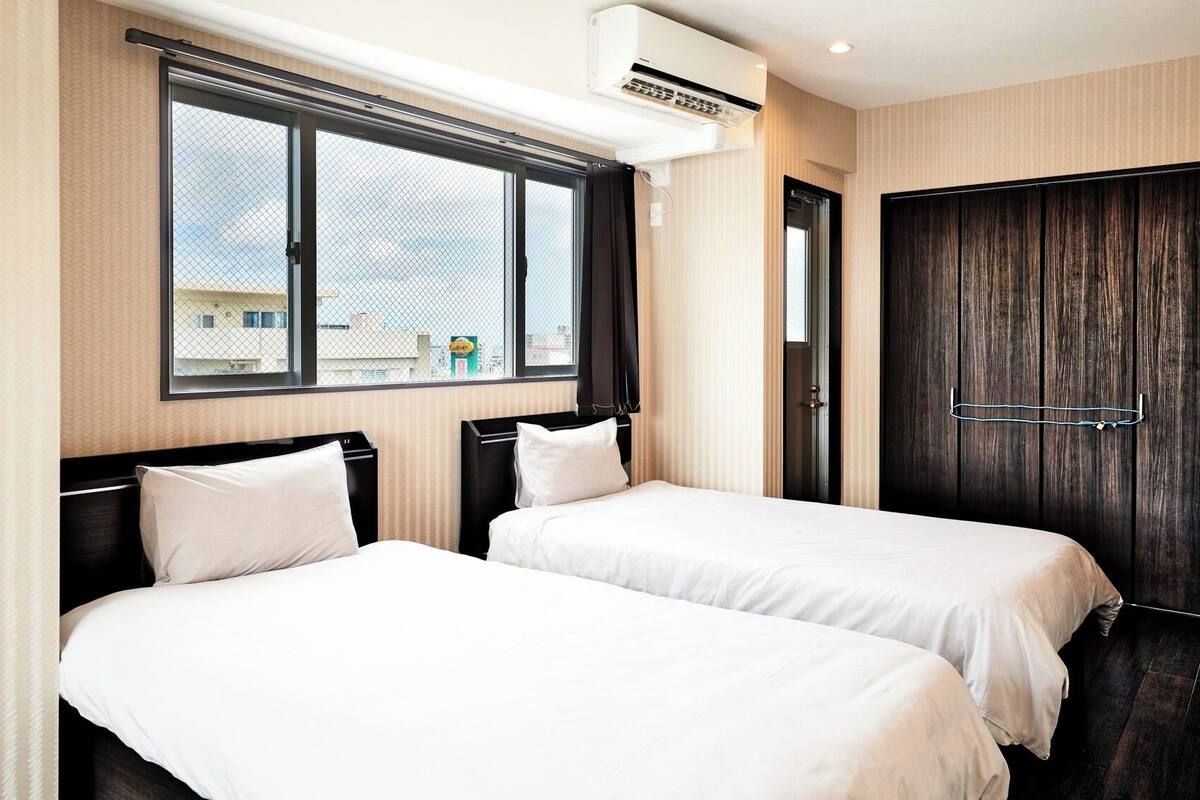 Une chambre d’hôtel avec deux lits, une grande fenêtre, un appareil de climatisation et une porte en bois foncé.