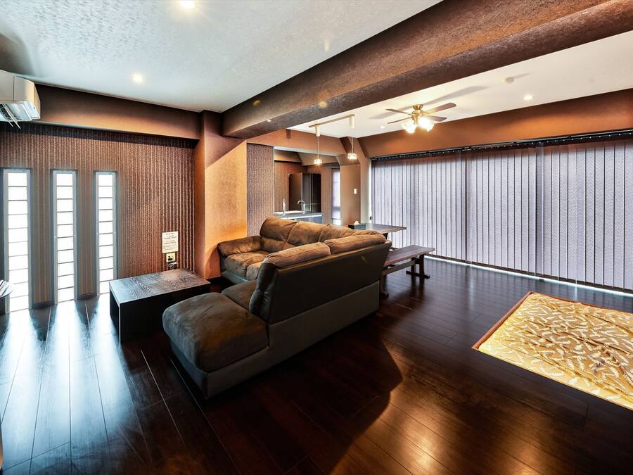 Una sala de estar moderna con un sofá, una mesa de centro y una mesa de comedor. Hay un ventanal con persianas, un ventilador de techo y un aire acondicionado instalado en la pared.