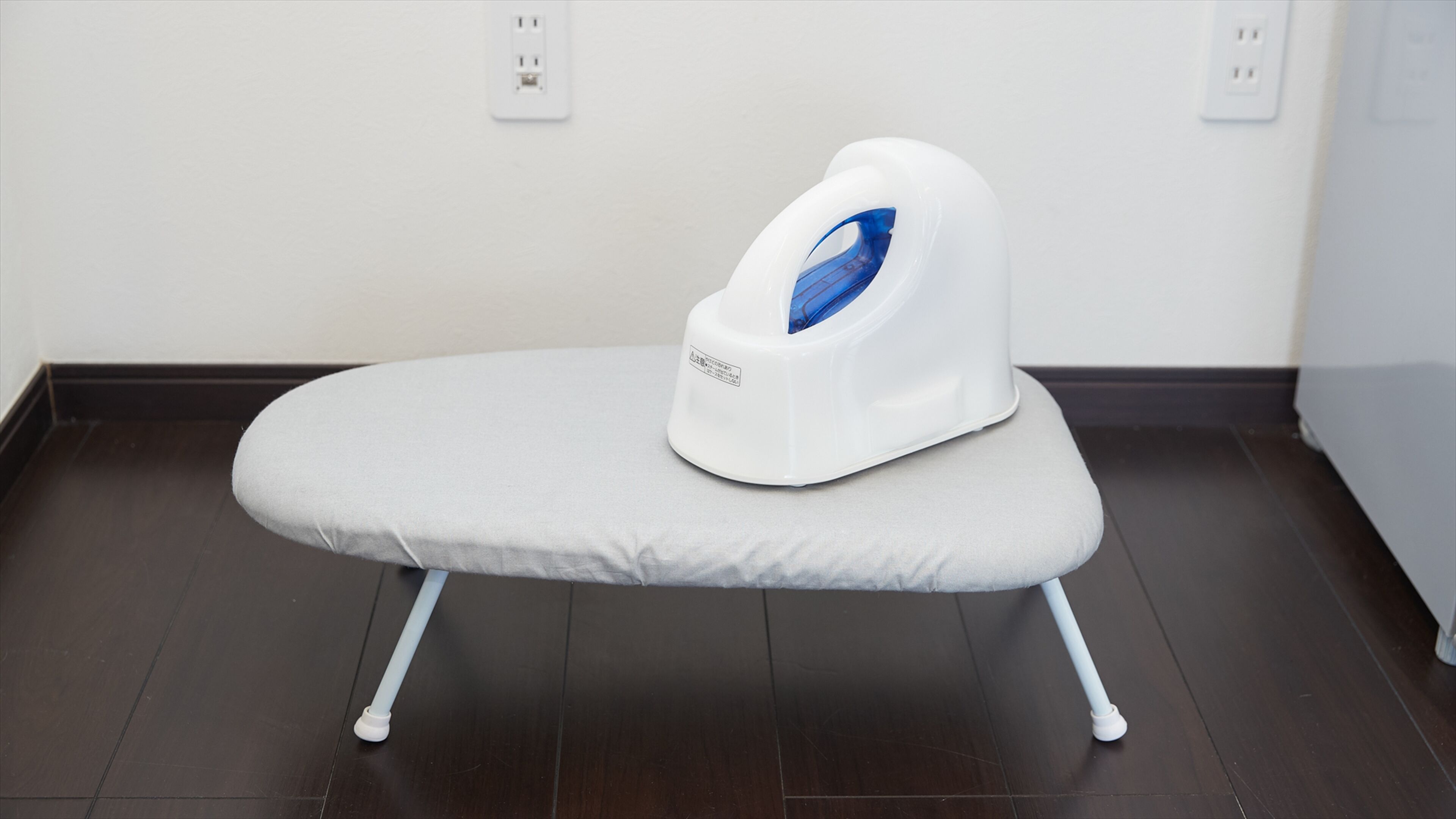 A white ironing board with a blue steam button, positioned on a dark wooden floor against a white wall with electrical outlets.