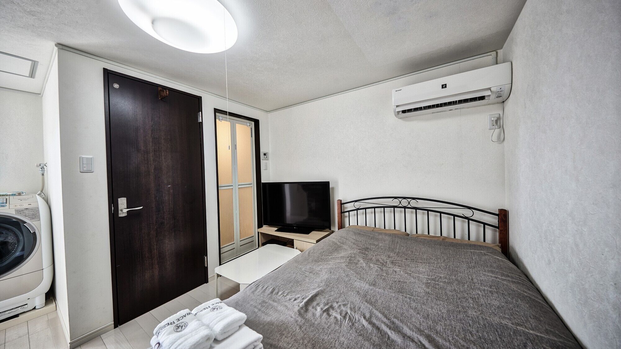 A bedroom with a bed, a TV on a stand, an air conditioner, a washing machine, and a door.