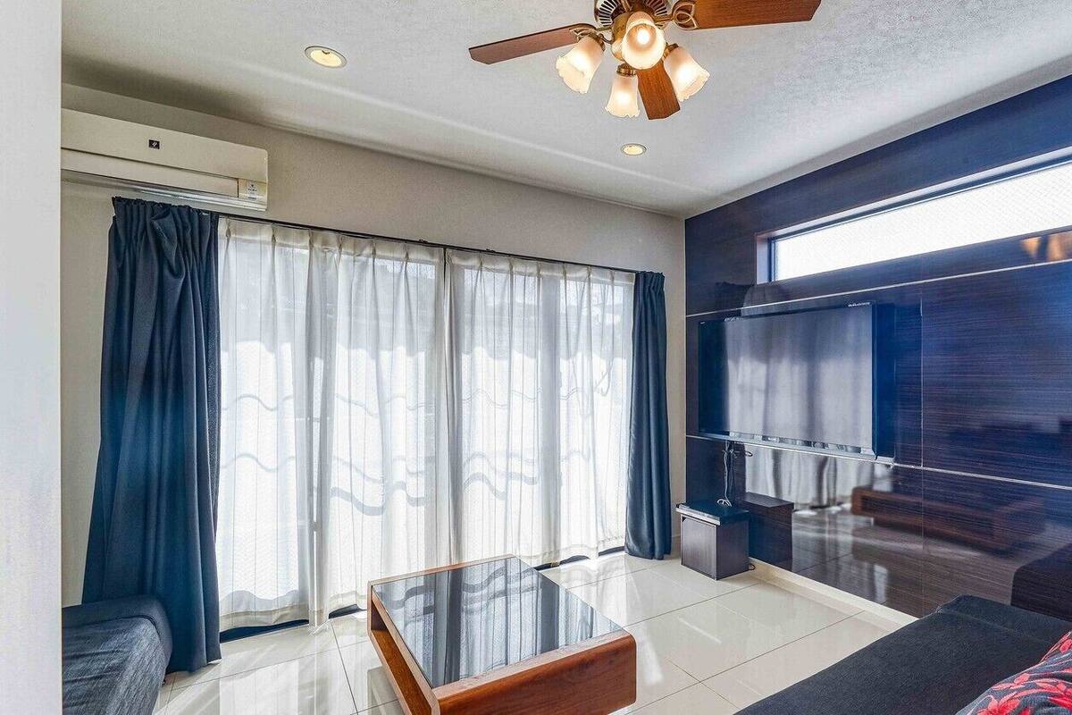 Una sala de estar moderna con una mesa de centro de vidrio, un sofá, un ventilador de techo y un ventanal con cortinas.