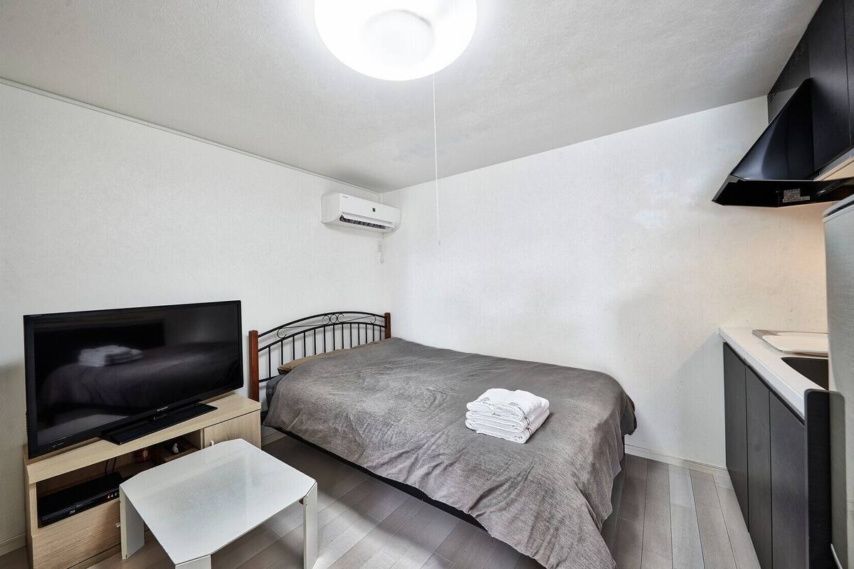 Una habitación compacta con cama, televisión, una mesita, un banco, kitchenette y luz de techo.