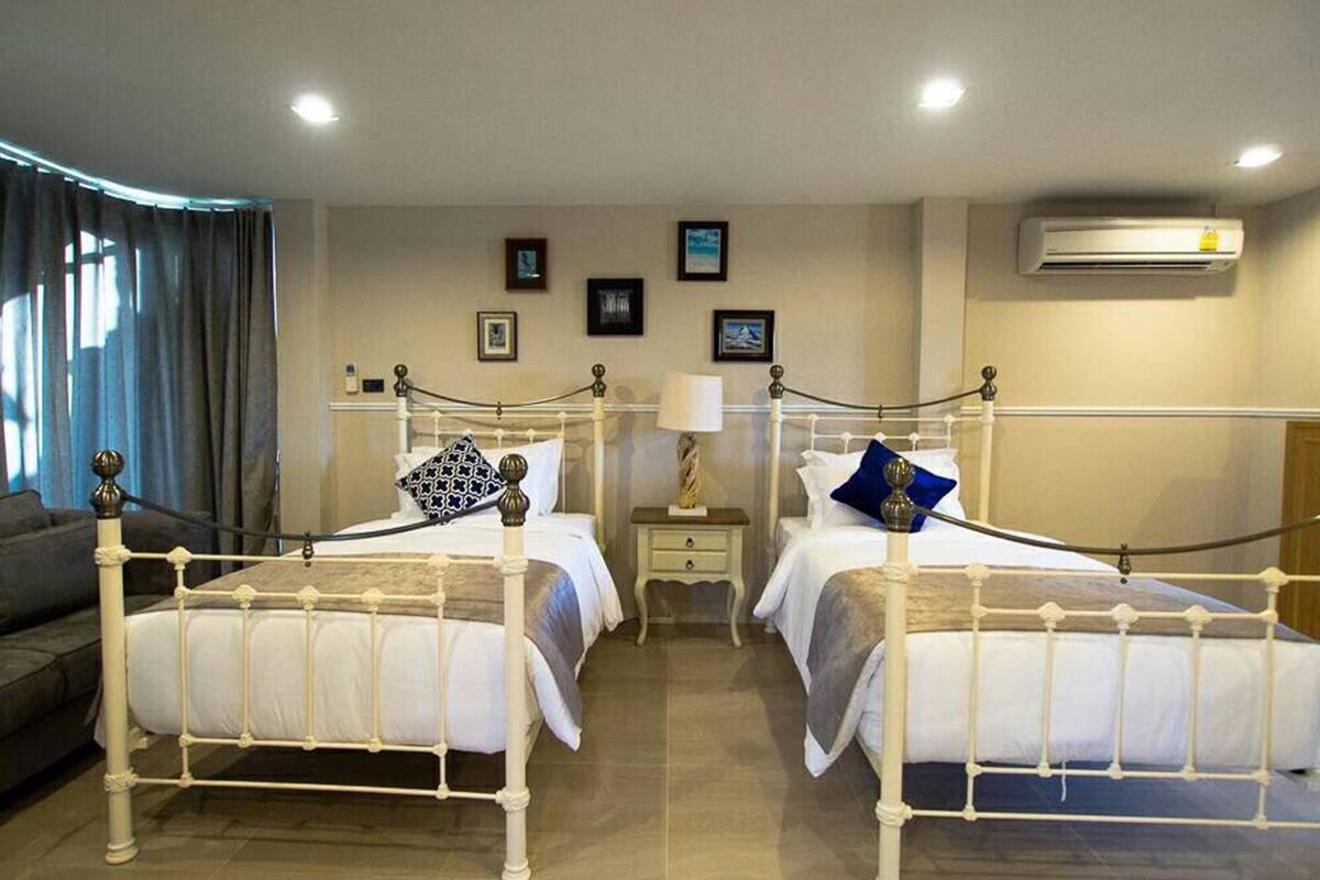 A hotel room with two white metal beds, a small bedside table with a lamp, a wall-mounted air conditioner, and a window with curtains.