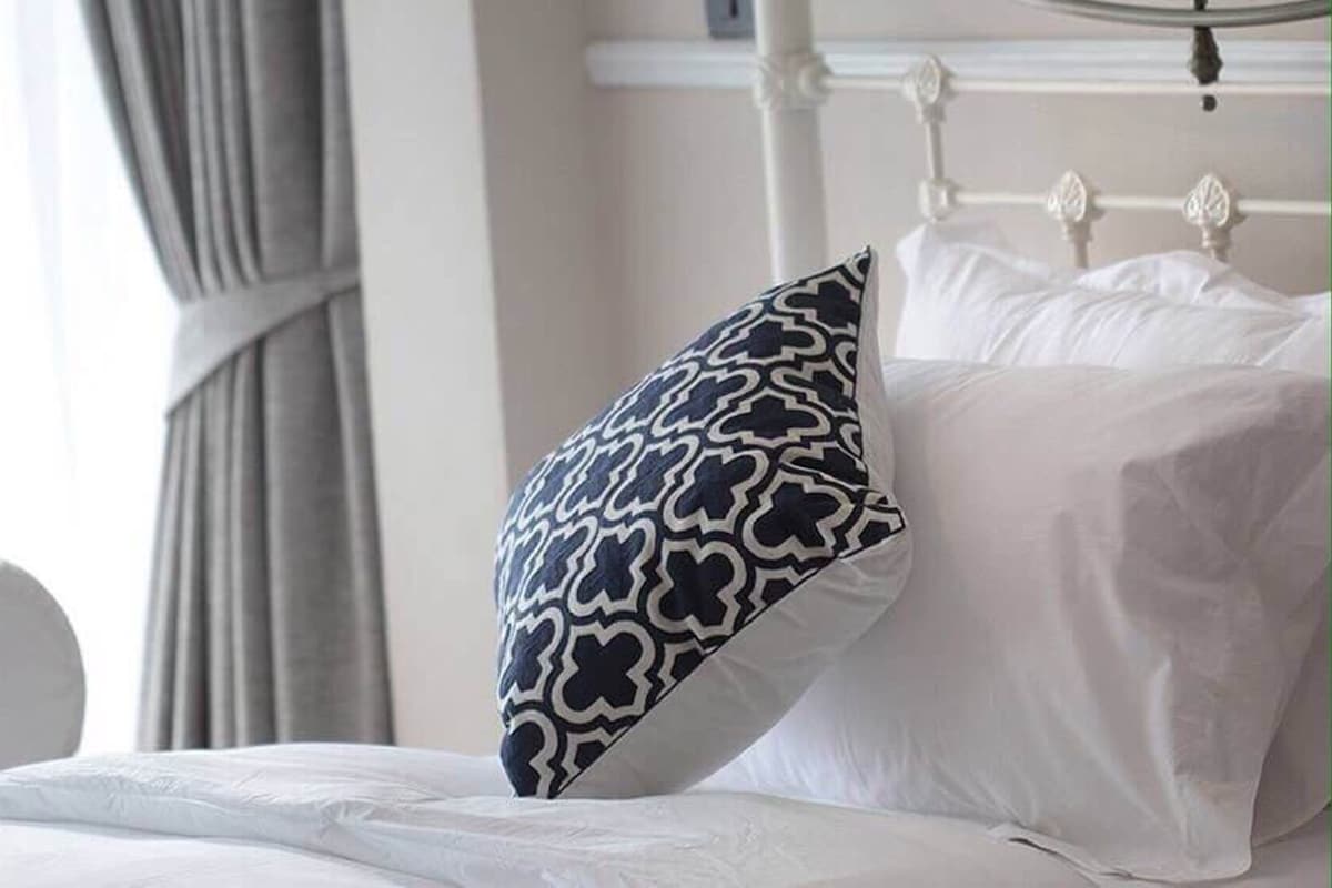 Una cama con ropa blanca y una almohada estampada, cabecera blanca y cortina.