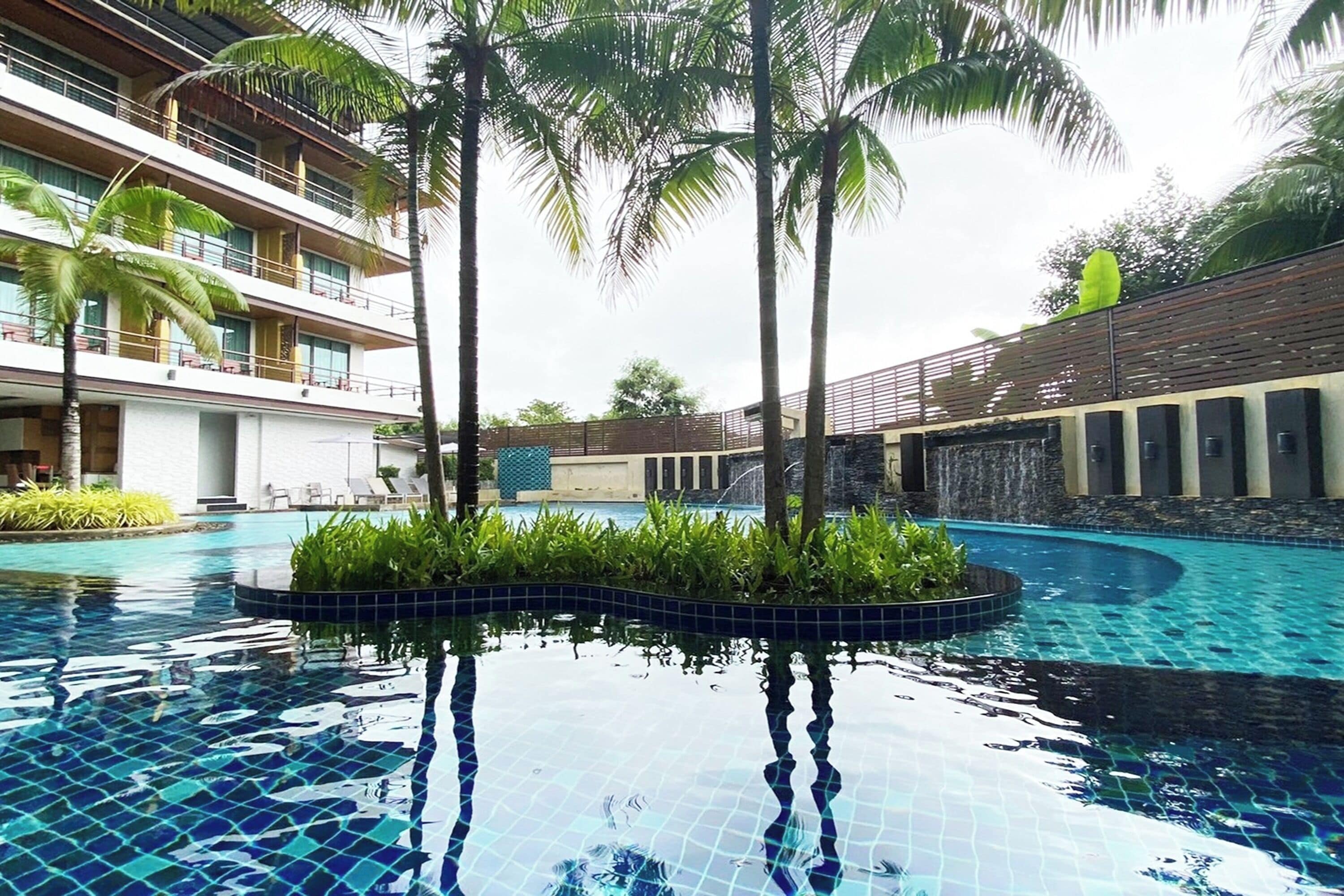 Ein Hotelpoolbereich mit klarem, blauem Wasser, Palmen und einem modernen Gebäude mit Balkonen.