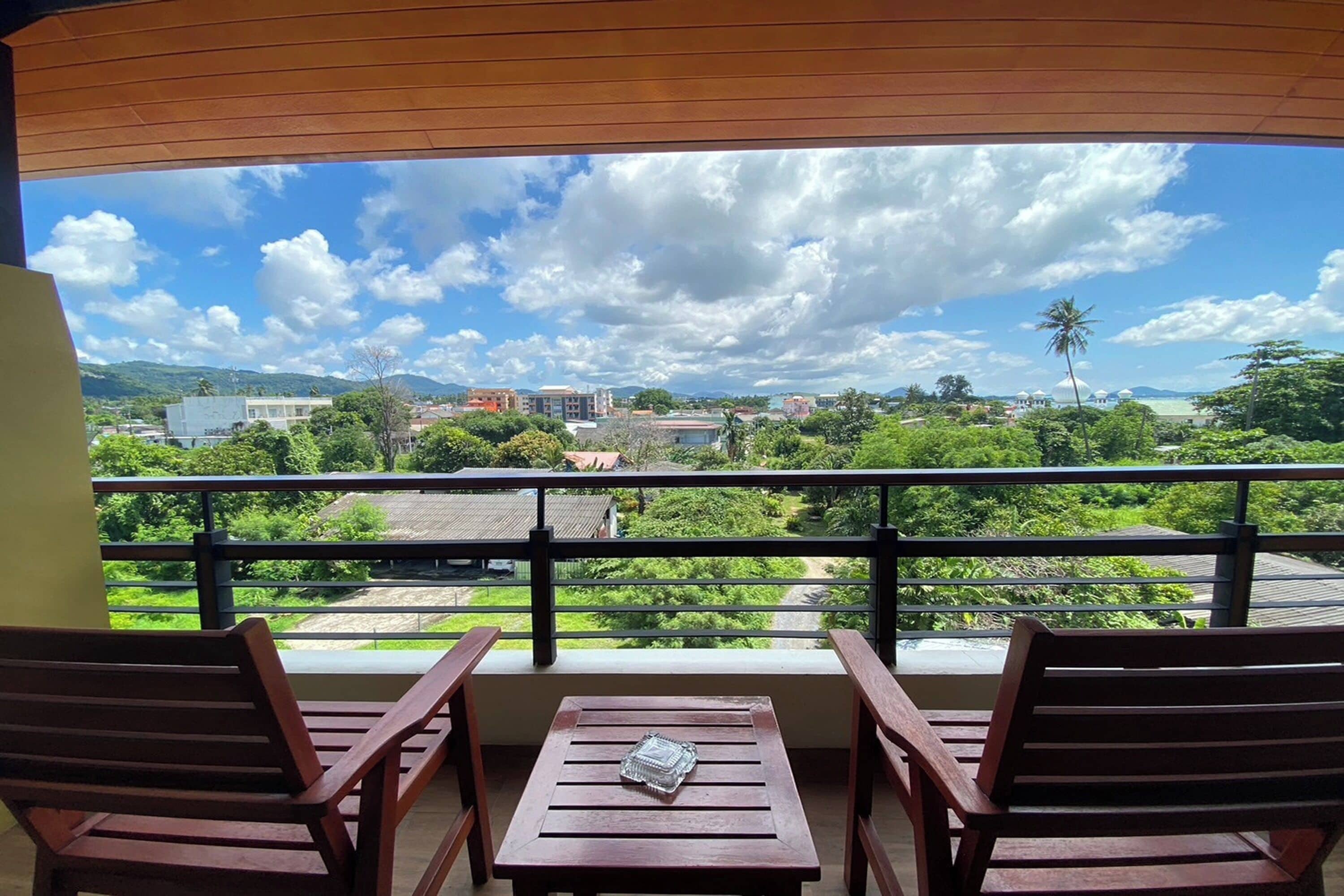 A balcony with two wooden chairs and a table, overlooking a scenic view of a beach, buildings, and greenery.