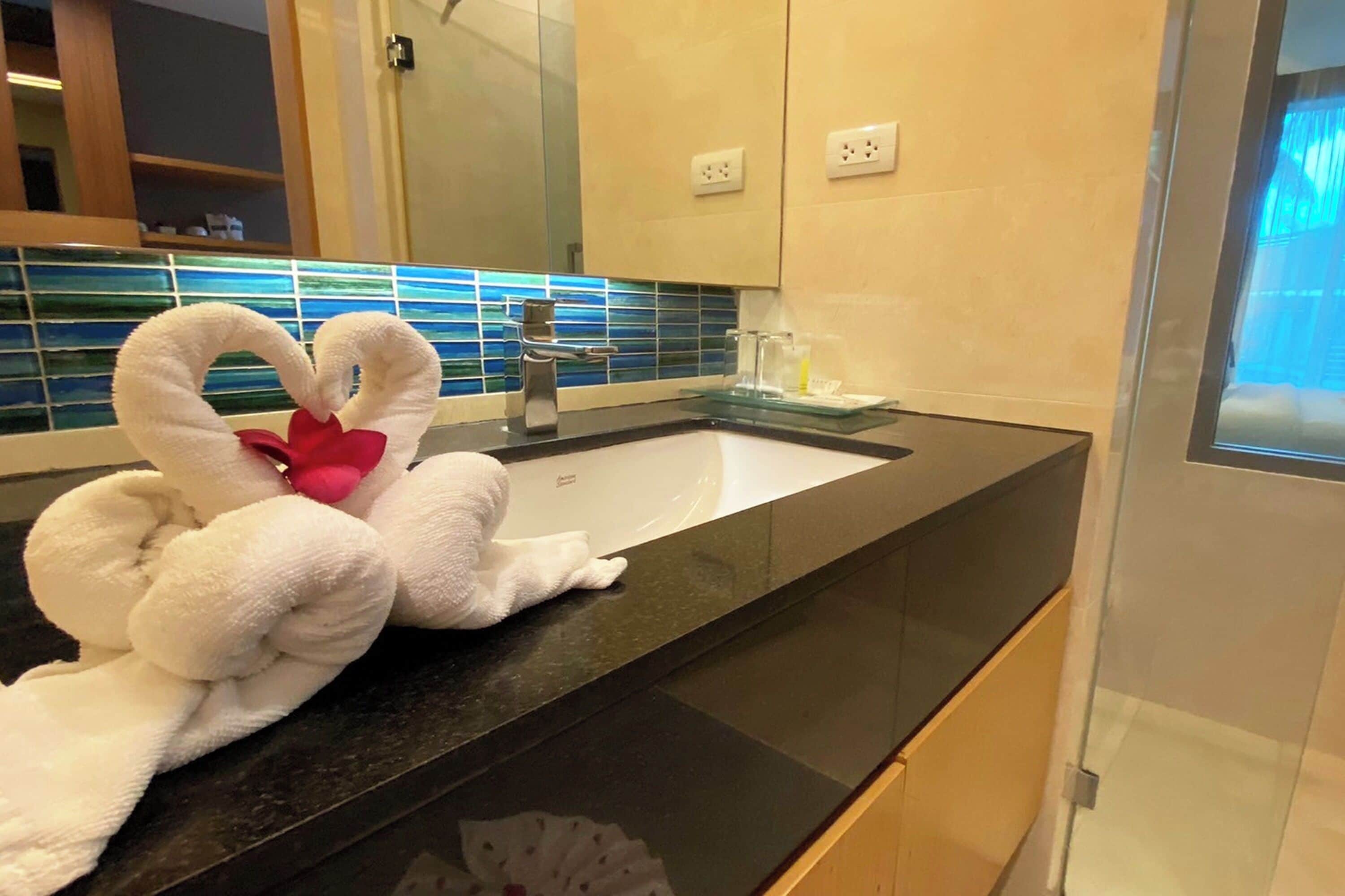 A bathroom with a black countertop, a white sink, and a mirror above it. There are two white towels shaped like swans on the countertop.