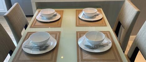 Une table à manger est dressée avec quatre bols et quatre cuillères blancs, posés sur des sets de table, et entourée de chaises.