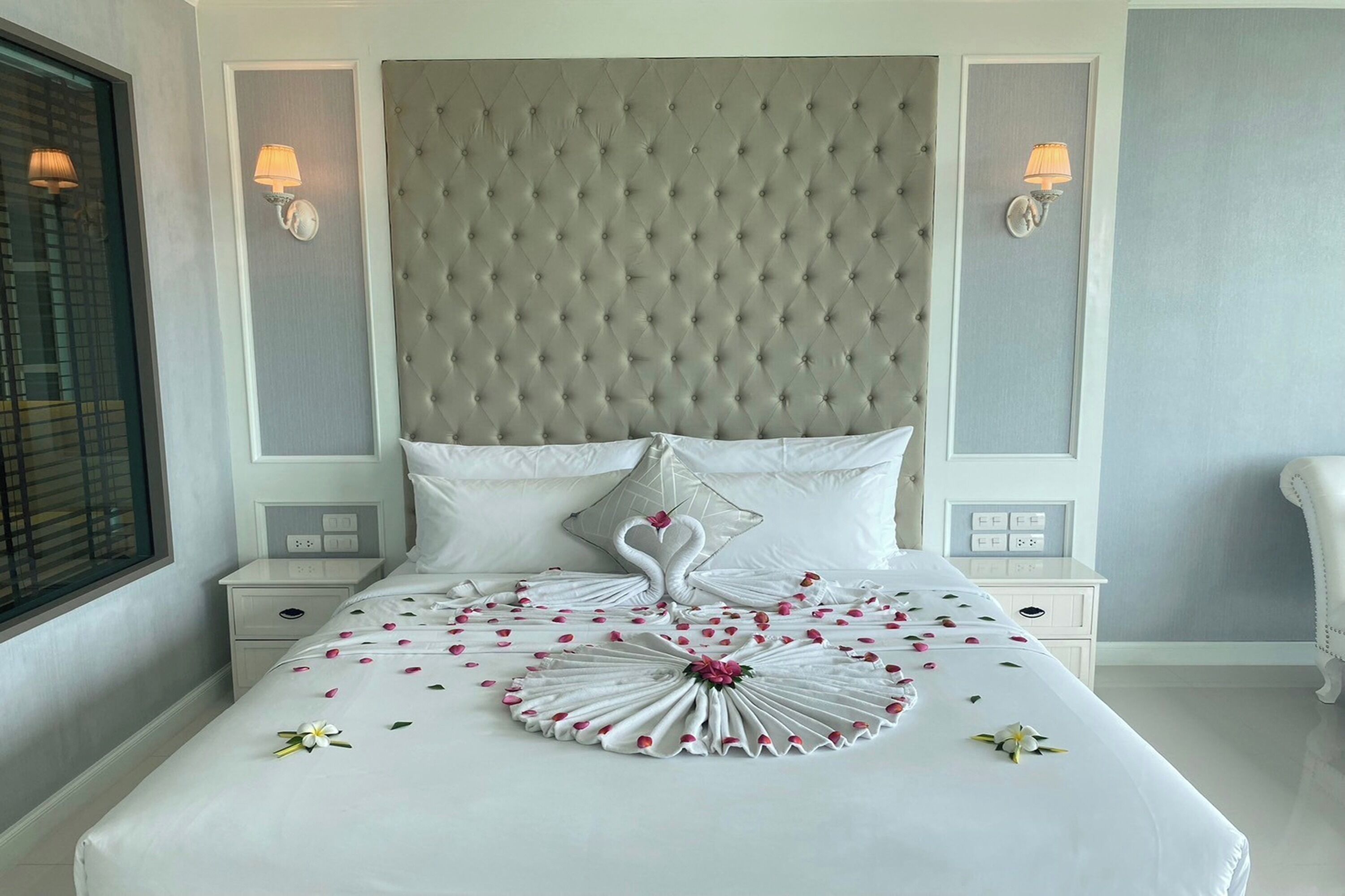 A neatly made bed with a swan design and rose petals, a tufted headboard, and bedside lamps.