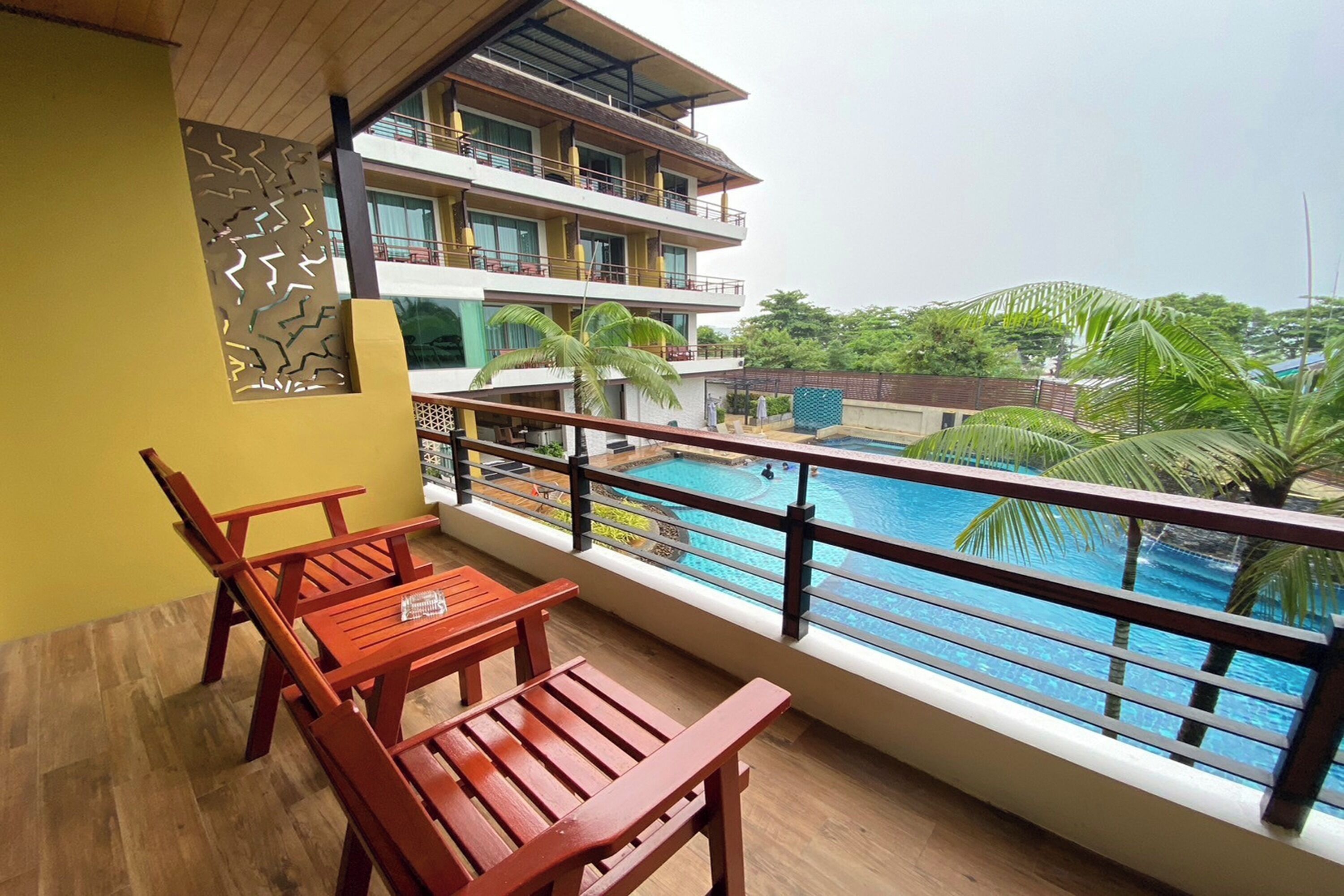 A balcony with wooden furniture overlooking a swimming pool and a multi-story building with balconies.