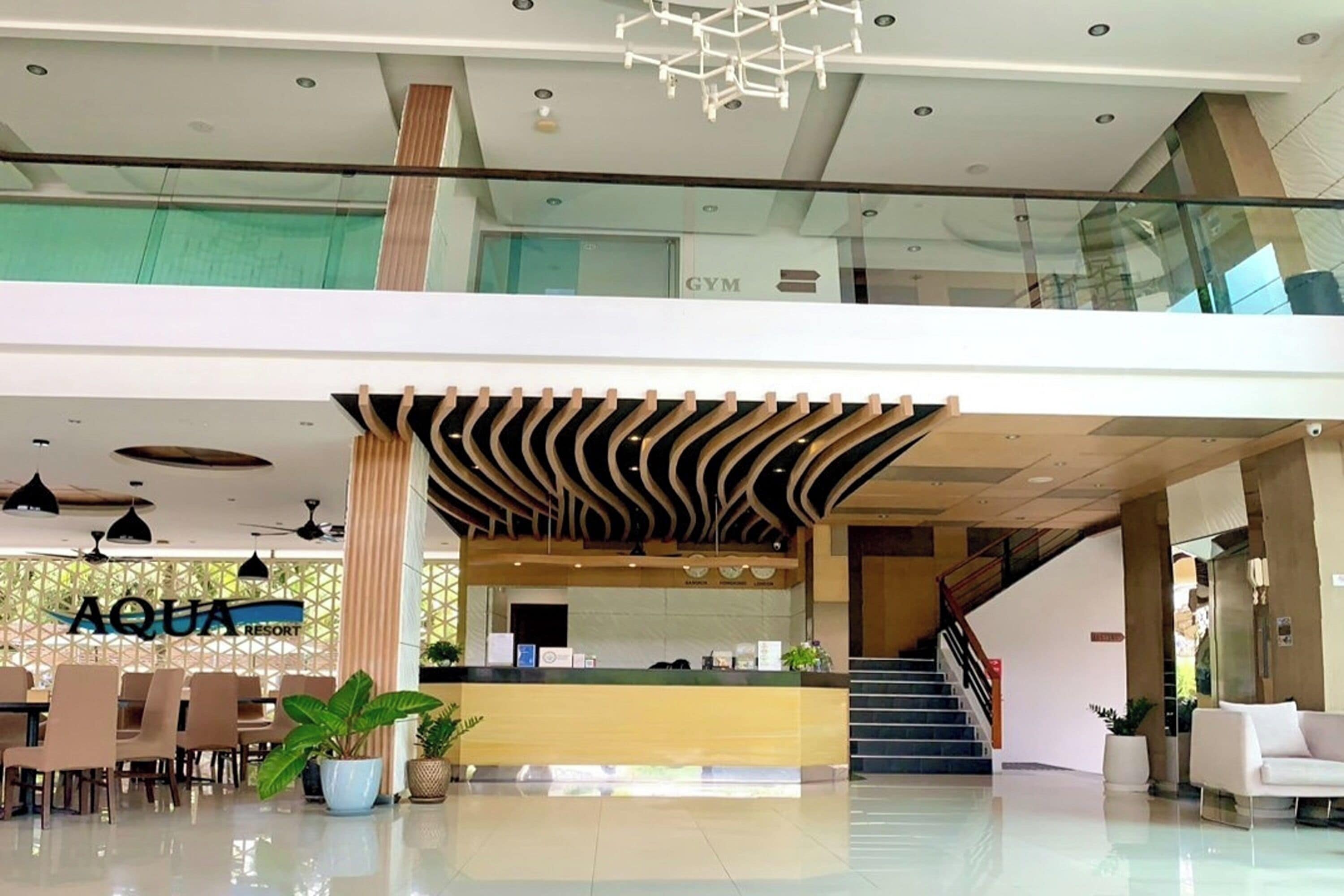 A modern hotel lobby with a reception desk, seating area, staircase, and a visible gym sign above.