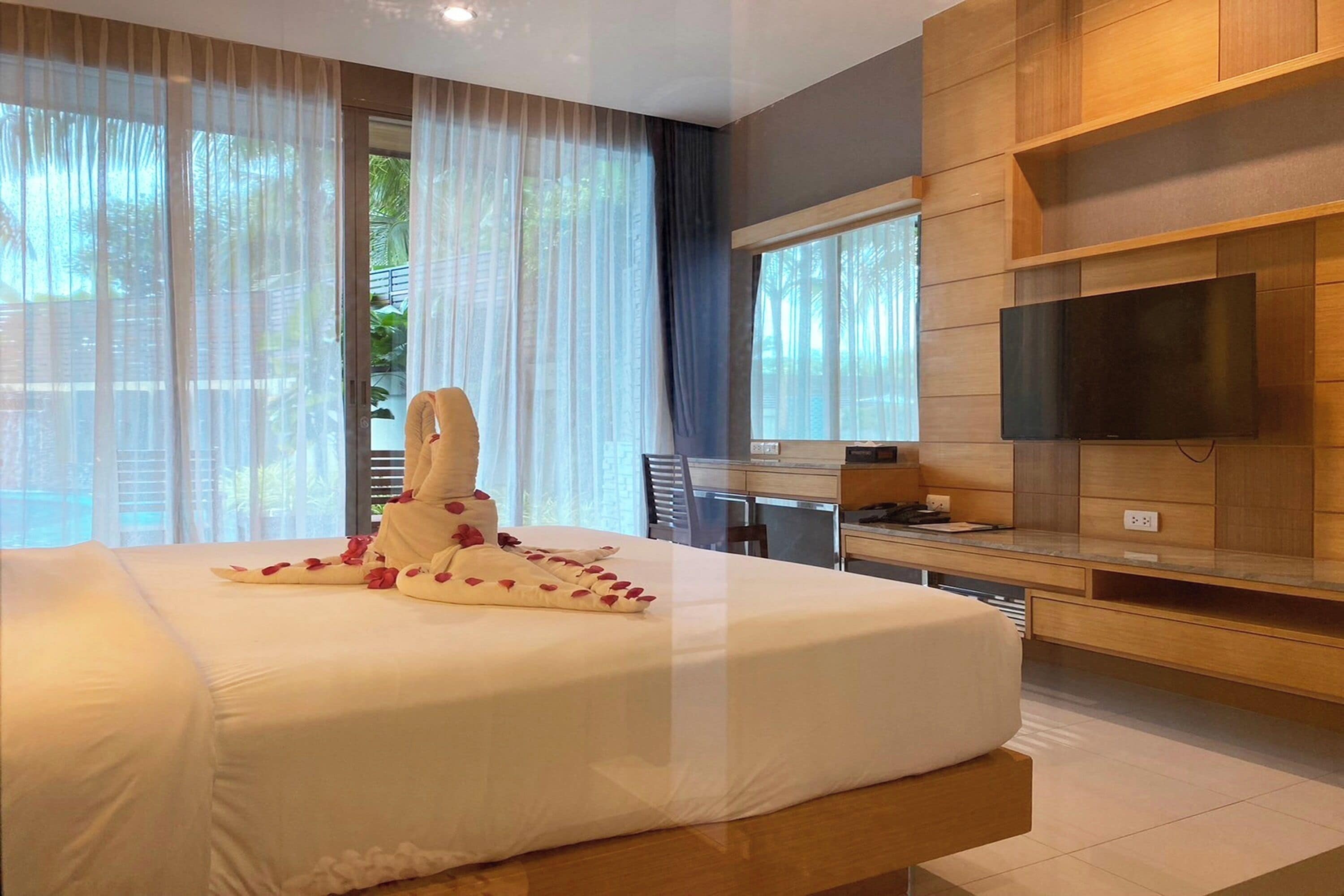 A modern hotel room with a large bed, a flat-screen TV mounted on the wall, and a view of greenery outside.