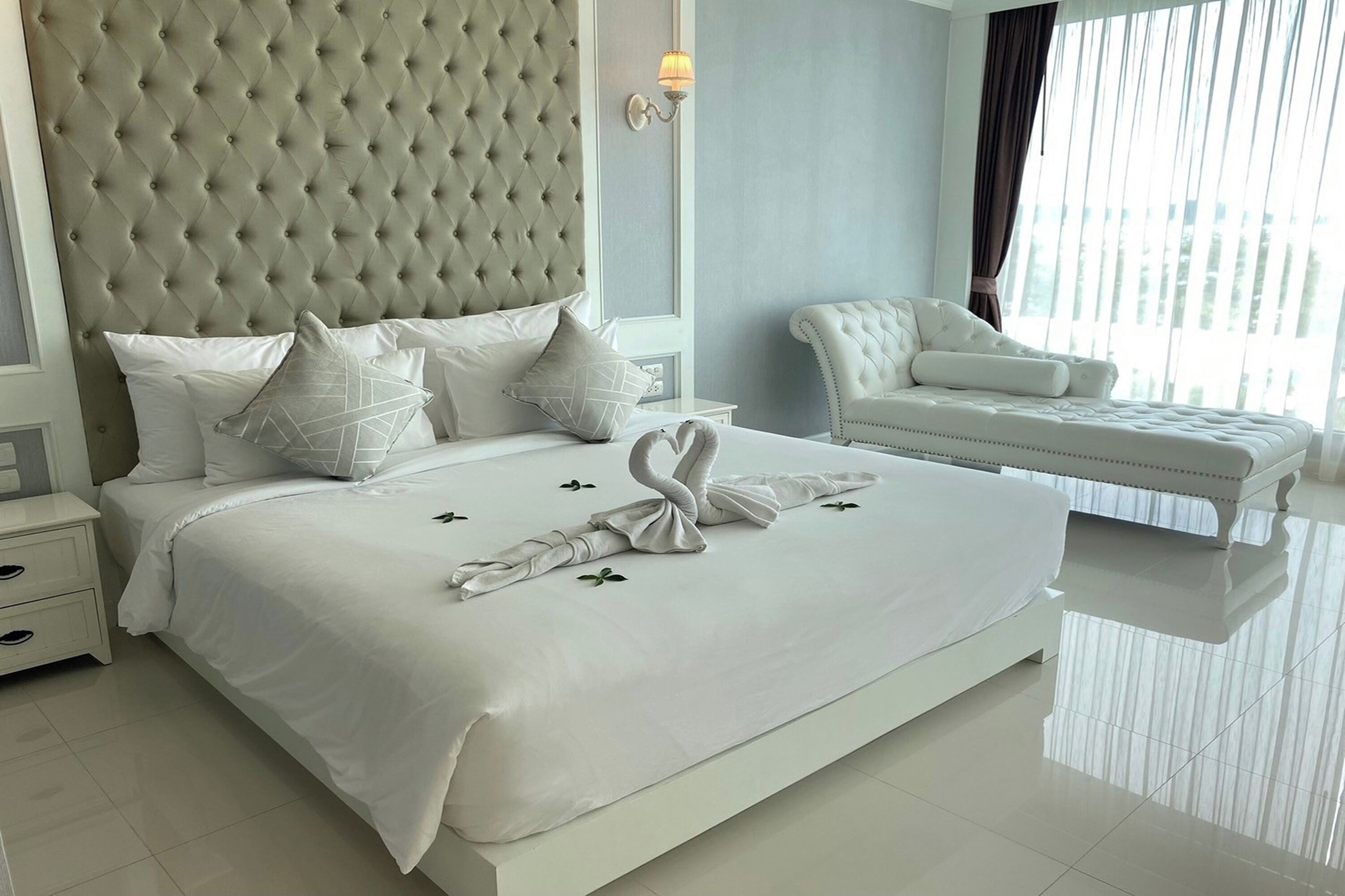 Un dormitorio moderno con un cabecero capitonado, una cama bien hecha con sábanas blancas y una escultura de toalla, un sillón blanco y un ventanal con cortinas.