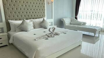 Un dormitorio moderno con un cabecero capitonado, una cama bien hecha con sábanas blancas y una escultura de toalla, un sillón blanco y un ventanal con cortinas.