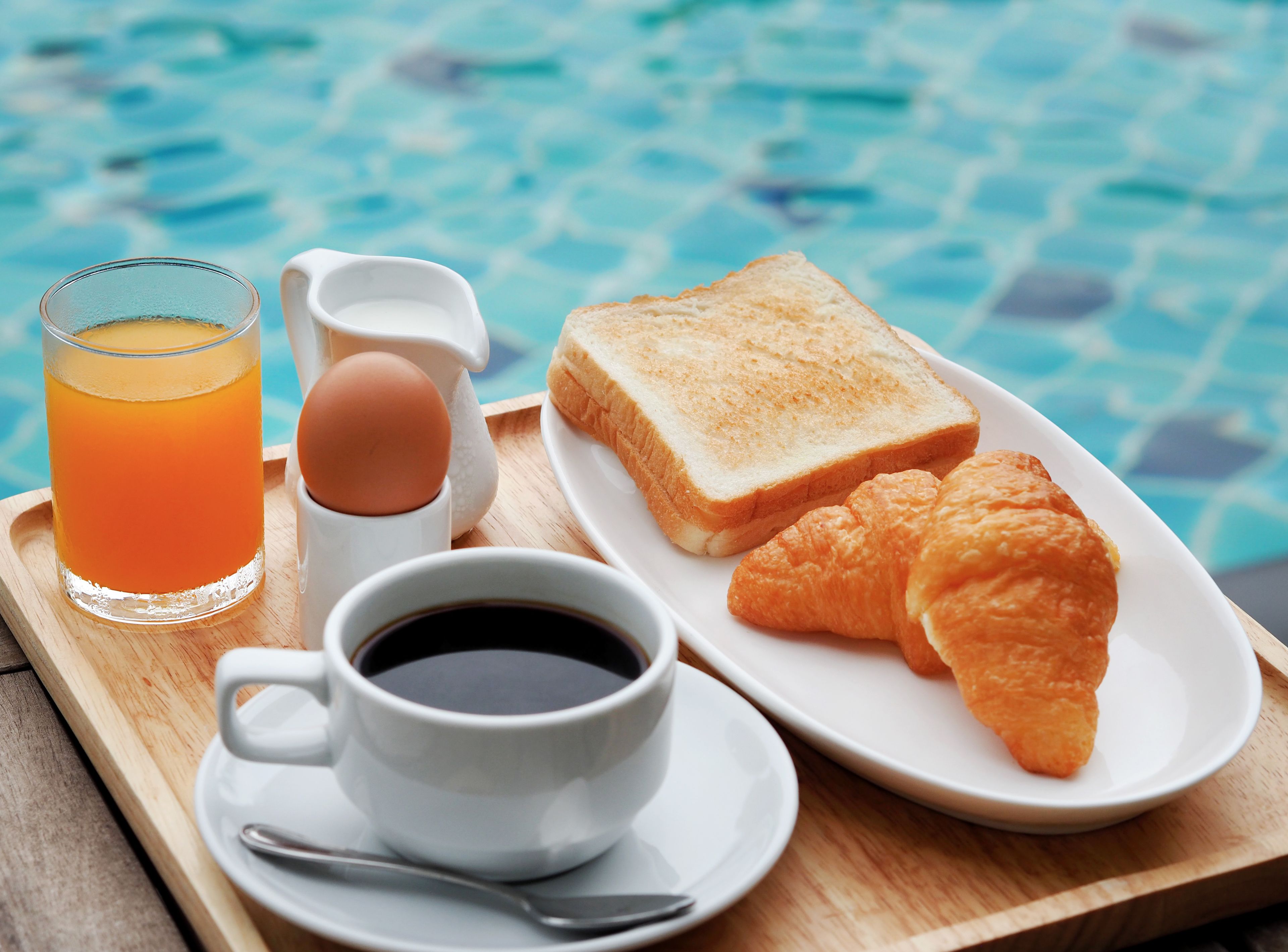 A breakfast tray with toast, croissants, a glass of orange juice, a cup of coffee, and a jug of milk by a pool.