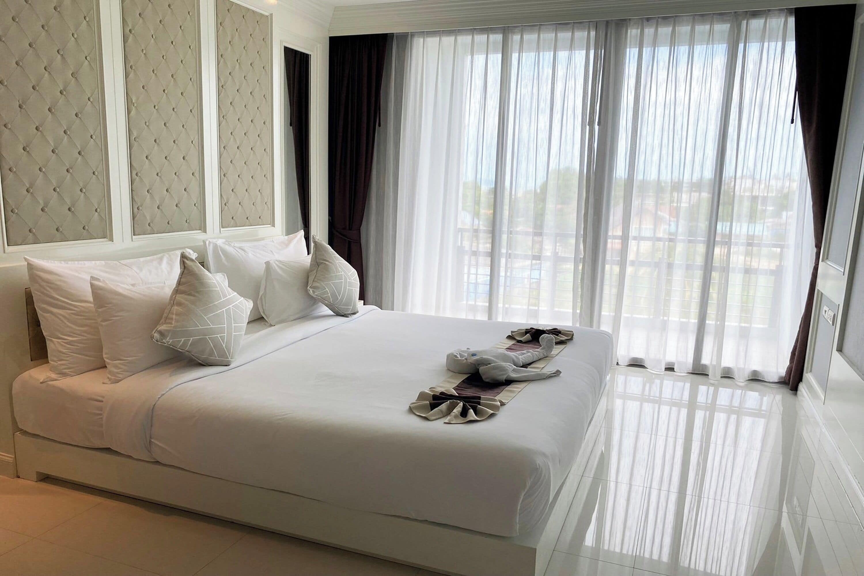 A hotel room with a large bed, white bedding, and a view through sheer curtains.