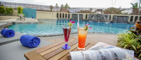 A wooden table with two drinks, a newspaper, and blue towels by a pool.