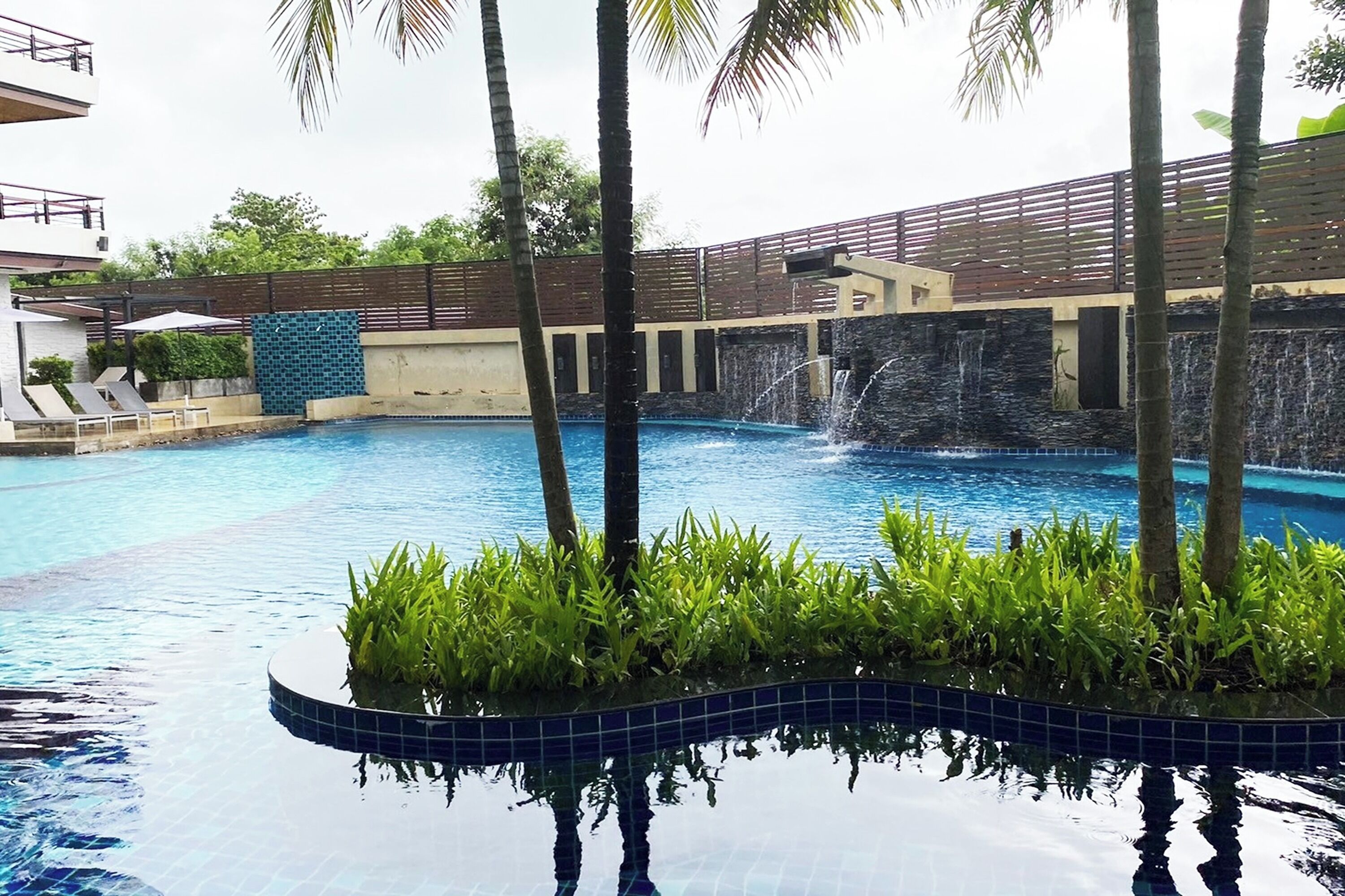 A swimming pool with a waterfall feature, surrounded by palm trees and a tiled edge.