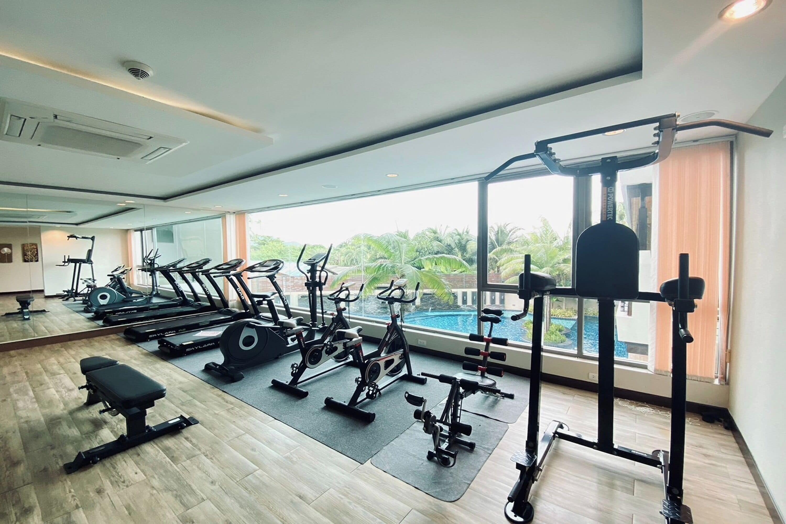 A gym with various exercise equipment, a bench, and large windows offering a view of a swimming pool and greenery.