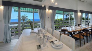 A dining area with a long table set for a meal, large windows with curtains, and a view of the ocean. - La Casa Panacea Okinawa Resort (Onna)
