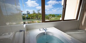 Un baño moderno con bañera, ventana con vista al océano y palmeras, y piso de baldosas.