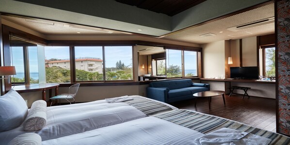 A hotel room with a large bed, a sofa, a small table, and a TV. The room has large windows offering a view of buildings and trees.