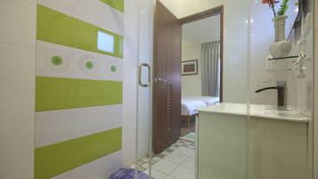 A bathroom with a shower, a sink, and a mirror. There is a door leading to a bedroom.